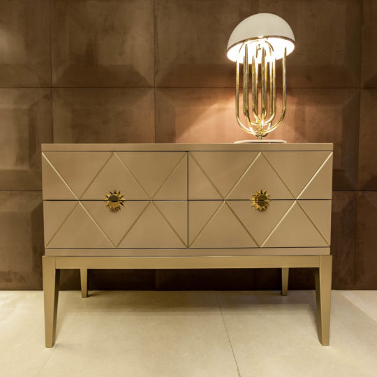 The Art Deco Inspired Chest of Drawers in beige features geometric diamond patterns, gold sunburst drawer handles, and a gold lamp with a white dome shade, set against a textured brown wall.
