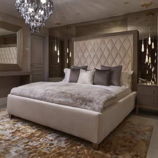 Luxurious bedroom with a large, plush Art Deco Inspired Upholstered Storage Bed featuring a tall headboard and numerous pillows. A modern chandelier hangs above, and pendant lights flank the bedside tables. The room is decorated in neutral tones with elegant textures and a large mirror.