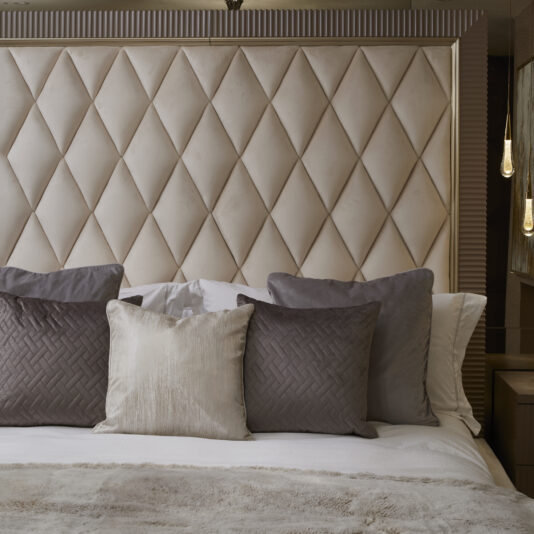 Art Deco Inspired Upholstered Storage Bed with Tall Headboard The Art Deco Inspired Upholstered Storage Bed with Tall Headboard features a diamond-tufted, Italian designer headboard and comes with four pillows: two charcoal grey with a herringbone pattern, one solid grey, and one beige. The bed is adorned with a beige blanket, complemented by two pendant lights hanging on either side.