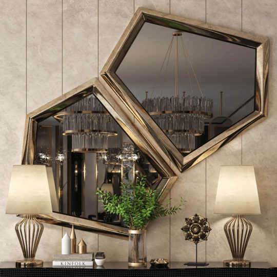 The Exclusive Pair Of Art Deco Inspired Mirrors with gold frames hang above a console table displaying modern lamps, a potted plant, sculpture, and a book; chandeliers reflect in the mirrors on a textured wall.
