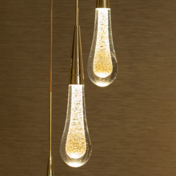 Set of 3 Glass Drop Pendant Lights A close-up of the Set of 3 Glass Drop Pendant Lights features elongated, teardrop-shaped bulbs filled with tiny sparkling elements that resemble glitter. The lights are illuminated, casting a warm and inviting glow against a textured, dark beige background.