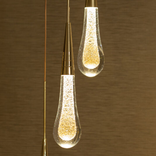 A close-up of the Set of 3 Glass Drop Pendant Lights features elongated, teardrop-shaped bulbs filled with tiny sparkling elements that resemble glitter. The lights are illuminated, casting a warm and inviting glow against a textured, dark beige background.
