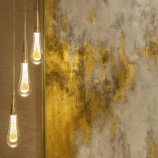A Set of 3 Glass Drop Pendant Lights with golden accents hang on the left side against a textured wall adorned with abstract gold and silver paint splashes, creating a modern and artistic ambiance.