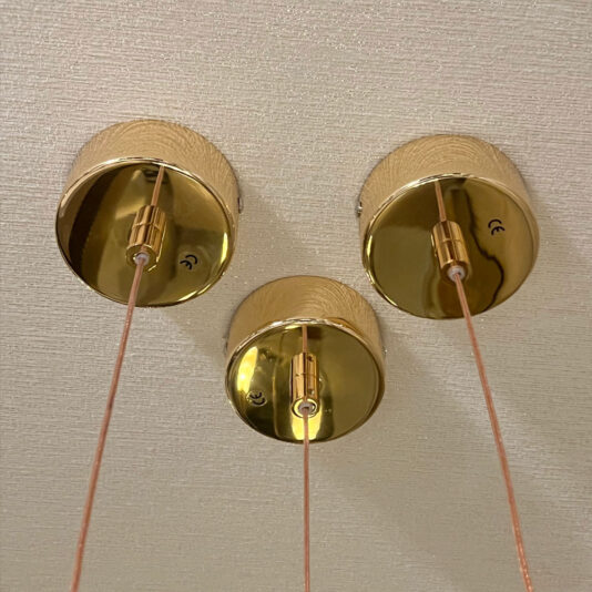 The Set of 3 Glass Drop Pendant Lights includes polished gold ceiling mounts with pink cords hanging from a beige ceiling. The circular, reflective mounts add an elegant touch to the pendant light fixtures.