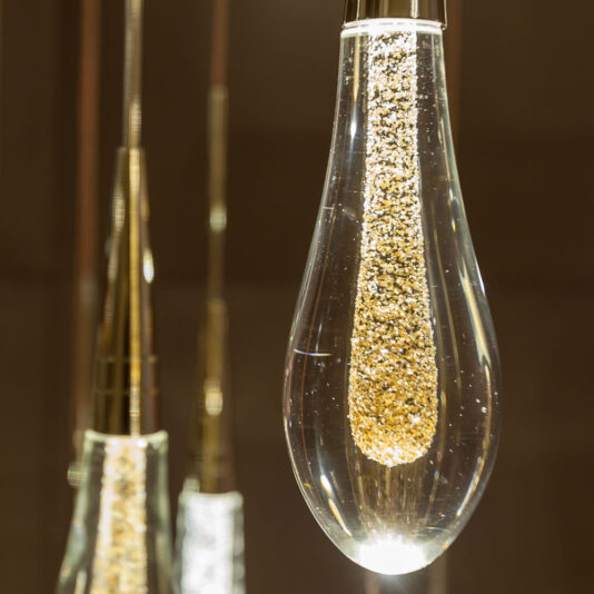 Close-up of an elegant teardrop-shaped pendant light fixture, filled with small, sparkling golden particles and emitting a warm glow. Several similar fixtures from the Set of 3 Glass Drop Pendant Lights are blurred in the background, suspended at various heights.