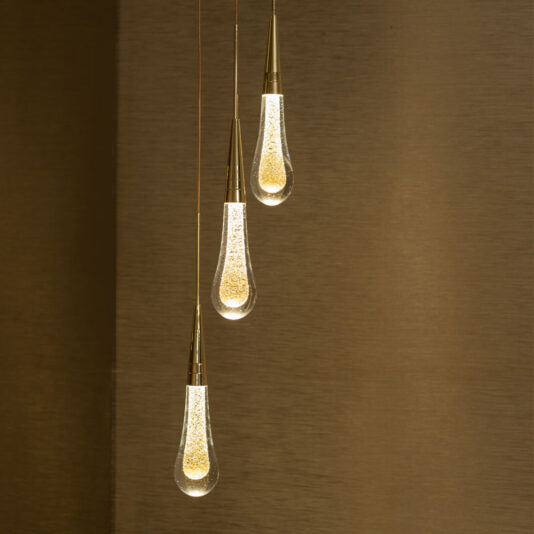 The Set of 3 Glass Drop Pendant Lights hang vertically in a row against a warm, brown background. Each teardrop-shaped light features a gold top and a clear lower part filled with small sparkles, creating an elegant and luxurious ambiance.
