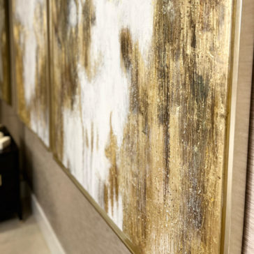 Original Gold Leaf Abstract Painting A close-up image showcases two Original Gold Leaf Abstract Paintings. The artwork displays a harmonious blend of white and gold hues with textured, streaky brushstrokes. Each painting is elegantly framed in a thin gold frame and hung on a beige wall, with part of a black piece of furniture visible nearby.
