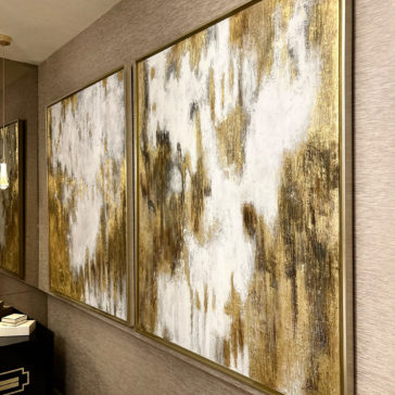 Original Gold Leaf Abstract Painting A hallway showcasing two Original Gold Leaf Abstract Paintings, featuring gold, white, and black textures on a beige wall. Both paintings are framed in gold and hanging side by side. Below them, a black console table displays a few stacked books and decorative items.