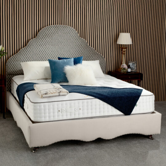 A modern bedroom with wood-panelled walls features a neatly made bed adorned with the Cawsand Ortho Mattress By Juliettes Interiors, a decorative headboard, layered pillows, a blue throw, a bedside table, lamp, and framed photos.