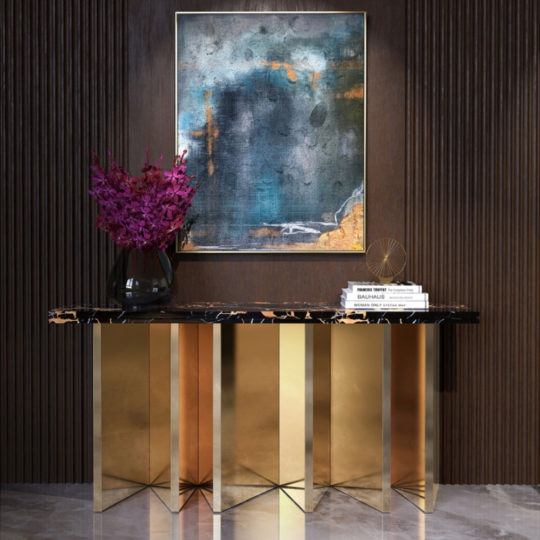 The Contemporary Marble Console with a black and gold marble top and gold geometric legs stands against a dark panelled wall, adorned with purple flowers in a vase, stacked books, and a sculpture beneath an abstract painting.