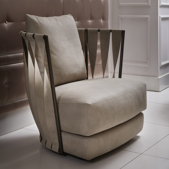 A Luxury Modern Leather Twist Armchair with a sleek, curved design is placed on a glossy white tile floor. The chair features wide beige cushions and metal accents along the sides, adding a contemporary touch to the space. The background includes part of a tufted wall panel and a white paneled wall.