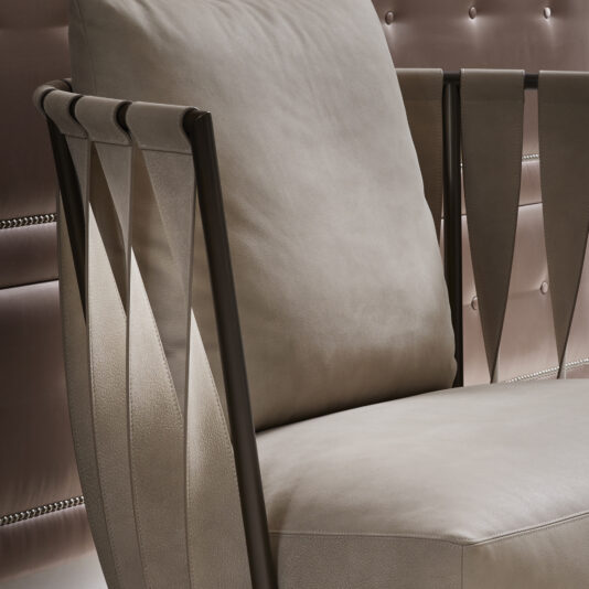 A close-up of the **Luxury Modern Leather Twist Armchair** showcases its beige leather seat and backrest. The armrests, featuring a contemporary twist, are made from arching dark metal frames with triangular leather panels. The chair is positioned against a quilted wall adorned with light brown cushioning and button details.