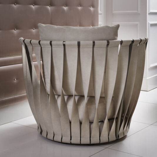 The Luxury Modern Leather Twist Armchair showcases a unique design with beige fabric and wide, vertical straps forming an open, rounded frame. Positioned in a contemporary room with white paneled walls, it features a cushioned, button-tufted surface detail.