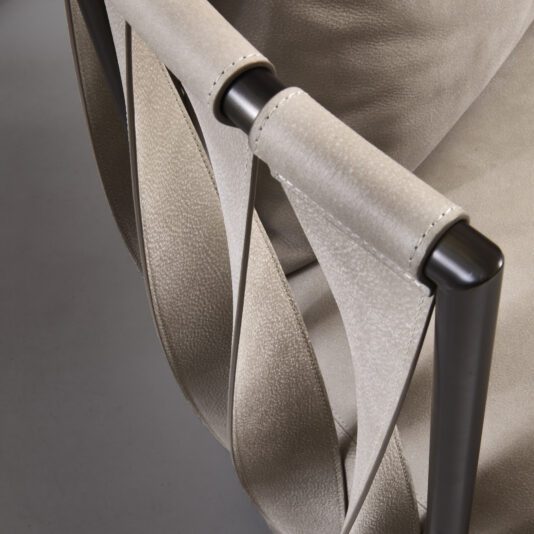 A close-up view of the Luxury Modern Leather Twist Armchair showcases its elegant beige upholstery and sleek metal frame. The chair's design is distinguished by unique twisted leather straps on the armrest, highlighting both aesthetics and craftsmanship. The background features a minimalist grey floor.