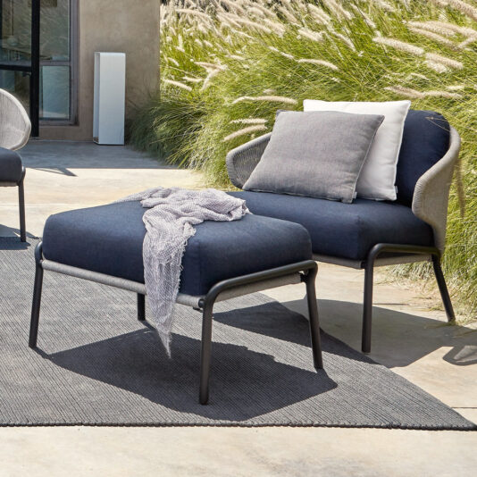 The Contemporary Outdoor Garden Armchair and Footstool feature dark cushions and a light throw, arranged on a grey rug near tall decorative grass on a sunny patio, with a cushion and neutral wall in the background.