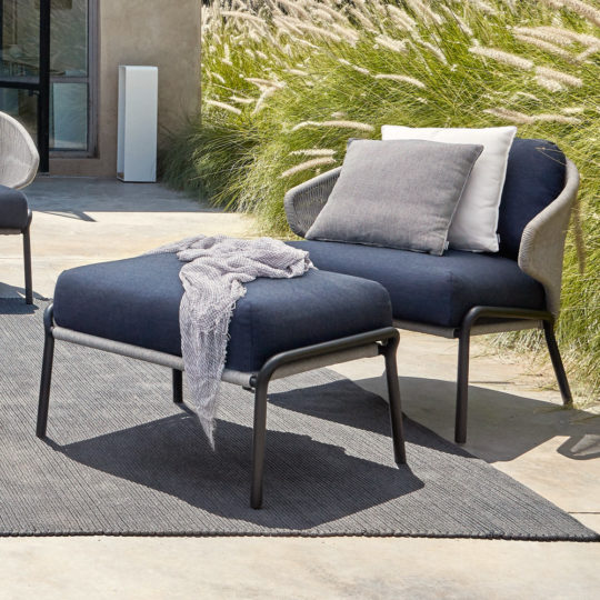 The Contemporary Outdoor Garden Armchair and Footstool feature dark cushions and a light throw, arranged on a grey rug near tall decorative grass on a sunny patio, with a cushion and neutral wall in the background.