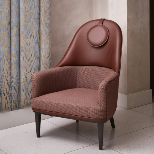 The High Backed Leather Occasional Armchair is a modern piece with a circular headrest, featuring a sophisticated brown leather upper section and a woven fabric lower section adorned with a subtle checkered pattern. The chair stands elegantly on dark wooden legs and is situated in a room complemented by patterned curtains.