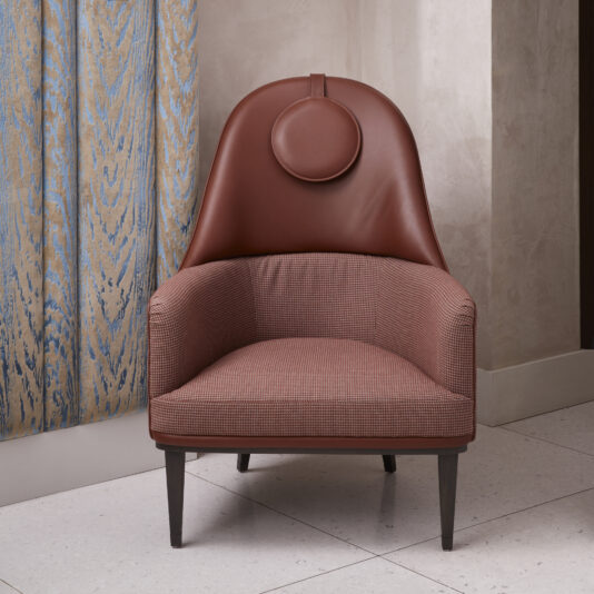 High Backed Leather Occasional Armchair The High Backed Leather Occasional Armchair, featuring a fabric upholstery, circular headrest, and dark wooden legs, is placed on a tiled floor next to a beige wall and patterned curtains. The chair boasts a modern, comfortable design.