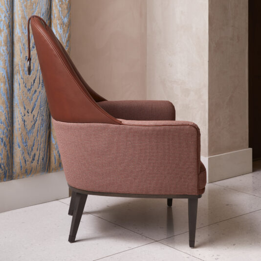 High Backed Leather Occasional Armchair The High Backed Leather Occasional Armchair, boasting a fabric seat and sides along with a high backrest, is positioned next to a patterned curtain in a neutral-toned room with light tile flooring. The chair features dark wooden legs and an elegant, modern design, epitomizing stylish comfort.