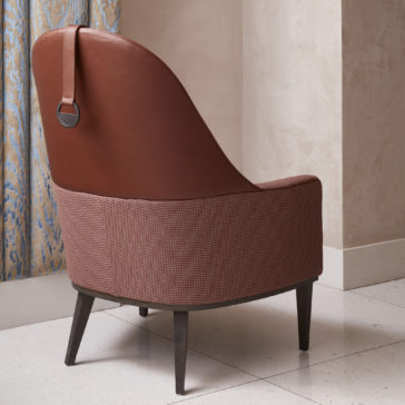High Backed Leather Occasional Armchair The High Backed Leather Occasional Armchair, featuring a brown upholstered lower half and dark wooden legs, is placed near a textured curtain. The armchair, which showcases a circular metal detail on the backrest, stands indoors on tiled flooring.