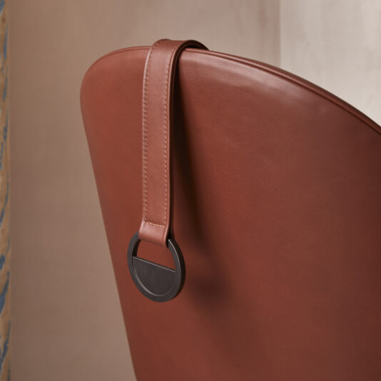 High Backed Leather Occasional Armchair Close-up of the backrest of the High Backed Leather Occasional Armchair. A decorative leather strap with a circular black and metal accent hangs gracefully over the top, adding an elegant touch to its modern design. The background is softly blurred.