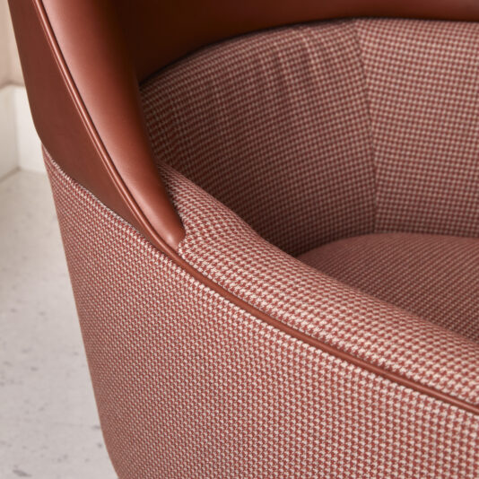 High Backed Leather Occasional Armchair A close-up view reveals the refined design of the High Backed Leather Occasional Armchair, showcasing a sophisticated brown leather upper portion paired with a stylish houndstooth-patterned lower fabric. The chair boasts smooth, clean lines and a dynamically textured fabric that artfully contrasts with the sleek leather. Enhancing its modern aesthetic is the light-colored, speckled floor beneath.
