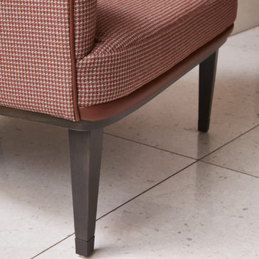High Backed Leather Occasional Armchair A close-up of a portion of the High Backed Leather Occasional Armchair with dark wooden legs and houndstooth-patterned upholstery in shades of red and cream shows the chair's elegant design. The light-colored tile floor beneath it features gray specks, adding to the sophisticated look.