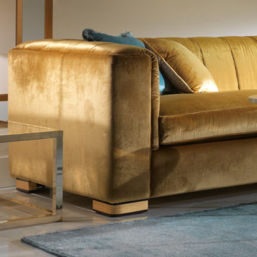 A luxurious High End Velvet 3 Seater Sofa in rich gold is showcased in a contemporary living room. The sofa is adorned with various cushions, and a light blue area rug is placed on the tiled floor in front. A sleek, metal side table stands to the left of the sofa. Cozy lighting enhances the scene.