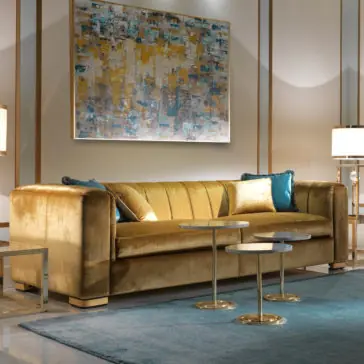 A luxurious living room featuring the High End Velvet 3 Seater Sofa in plush gold, adorned with blue and gold cushions. Two modern, round, gold side tables with marble tops sit in front of it. On the wall hangs an abstract painting with shades of blue, gold, and white. A blue rug graces the floor.