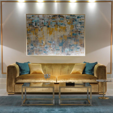 A modern living room showcases a High End Velvet 3 Seater Sofa in gold, adorned with teal and gold cushions. Above the sofa, an abstract painting displays hues of blue, gold, and white. In front of the sofa are two nesting tables holding decorative items over a teal rug.