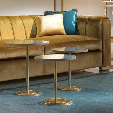 A modern living room scene features a High End Velvet 3 Seater Sofa adorned in yellow, complemented by gold and teal pillows. In front of the sofa, three round marble-top side tables with gold bases are arranged elegantly on a blue rug.