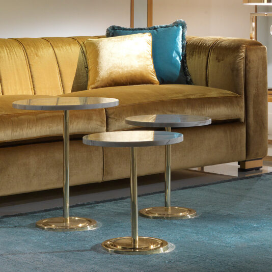 High End Velvet 3 Seater Sofa A modern living room scene features a High End Velvet 3 Seater Sofa adorned in yellow, complemented by gold and teal pillows. In front of the sofa, three round marble-top side tables with gold bases are arranged elegantly on a blue rug.