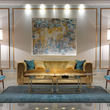 A stylish living room showcases a High End Velvet 3 Seater Sofa in yellow, adorned with blue and beige cushions, set on a blue area rug. In front of the sofa are two glass-top coffee tables. An abstract painting hangs above the sofa, with wall lights and modern lamps illuminating the space.