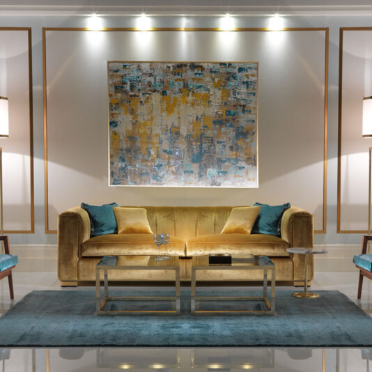 High End Velvet 3 Seater Sofa A stylish living room showcases a High End Velvet 3 Seater Sofa in yellow, adorned with blue and beige cushions, set on a blue area rug. In front of the sofa are two glass-top coffee tables. An abstract painting hangs above the sofa, with wall lights and modern lamps illuminating the space.