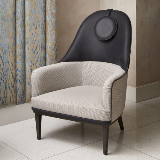The Modern High Backed Occasional Armchair is a contemporary piece featuring black leather and light-colored patterned upholstery. It boasts a tall, arched backrest with a circular black headrest and stands elegantly on four tapered legs. This stylish chair is set on a tiled floor next to a blue and gold patterned curtain.