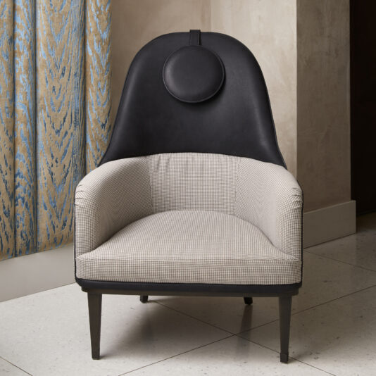 Modern High Backed Occasional Armchair The Modern High Backed Occasional Armchair features a checkered fabric seat and backrest, complemented by a black leather upper backrest and a circular leather accent at the top. It stands on four black legs and is placed on a light-colored tile floor near textured drapes.