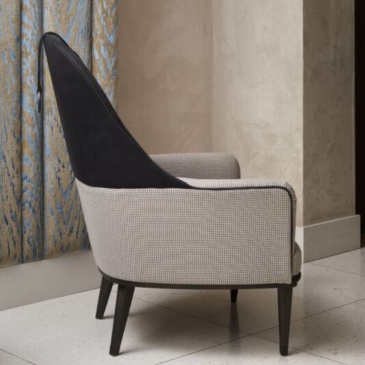 Modern High Backed Occasional Armchair A Modern High Backed Occasional Armchair, featuring a high backrest upholstered in gray fabric with black accents, is positioned in front of a wall adorned with light beige textured wallpaper. The armchair's slim, angled black legs complement its modern design and it sits next to a blue and beige patterned curtain.