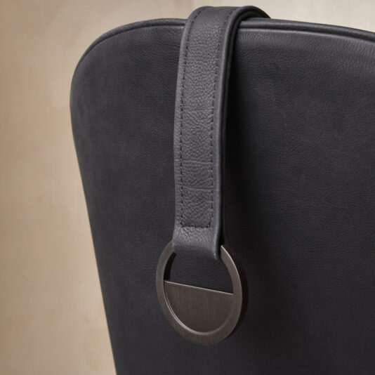 Modern High Backed Occasional Armchair Close-up of the Modern High Backed Occasional Armchair's black leather chair back, showcasing a sleek, metallic ring attached to a leather strap elegantly draped over the top. The chair is set against a blurred, neutral-toned wall.