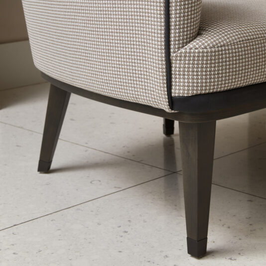 Modern High Backed Occasional Armchair A close-up of the bottom part of the Modern High Backed Occasional Armchair, showcasing its houndstooth upholstery. The chair features wooden legs with a dark finish, and is situated on a light-colored tiled floor, with one leg partially visible.