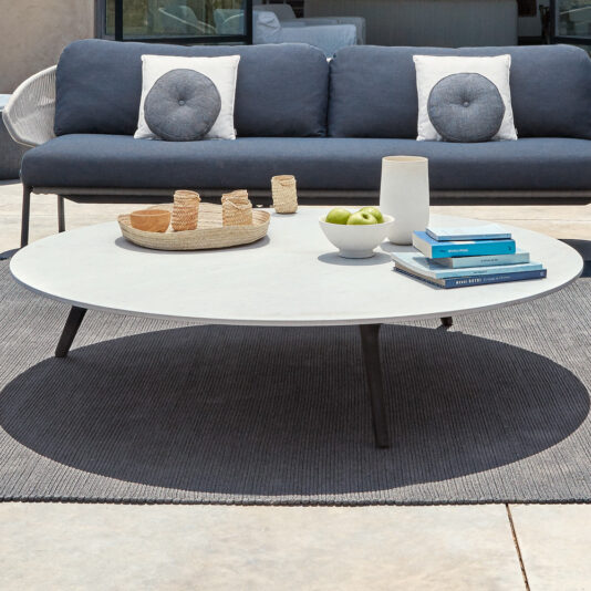A Modern Round Outdoor Coffee Table sits on a grey rug in front of a dark grey sofa with round, buttoned cushions on the patio. Baskets, apples in a bowl, books, and a vase are arranged on the table.