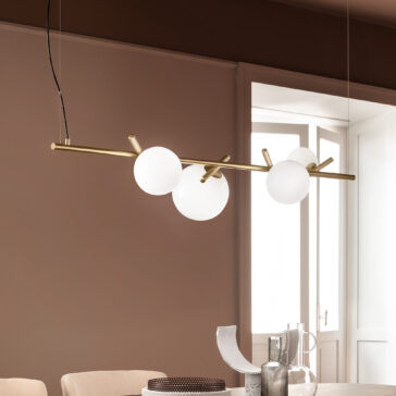 The Contemporary Murano Glass Horizontal Chandelier, featuring three frosted globes on a brass rod, elegantly hangs in a dining room with beige walls. Below the chandelier sits a table adorned with decorative items such as a silver pitcher, a round textured placemat, and an abstract art object.