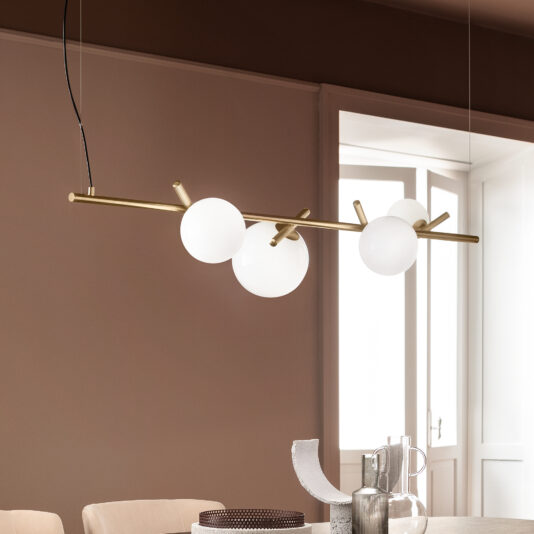 The Contemporary Murano Glass Horizontal Chandelier, featuring three frosted globes on a brass rod, elegantly hangs in a dining room with beige walls. Below the chandelier sits a table adorned with decorative items such as a silver pitcher, a round textured placemat, and an abstract art object.