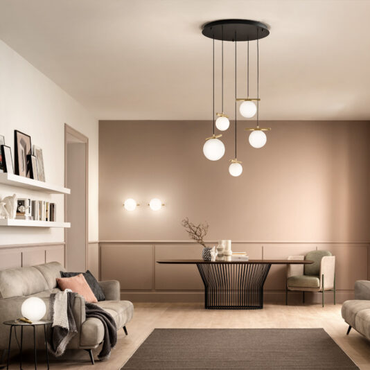 A modern living room with beige walls and wooden flooring. It features a gray sofa, a black wireframe coffee table, a green chair, and decorative items on shelves. A stylish Contemporary Murano Glass Pendant Light Chandelier with spherical bulbs hangs from the ceiling, and wall sconces add ambient lighting.