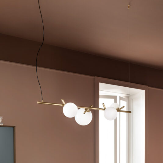 The Contemporary Murano Glass Horizontal Chandelier, featuring a sleek golden rod and three Murano glass globe-shaped bulbs, is suspended from the ceiling by two thin wires. Positioned in a room with brown walls and a large window letting in natural light, this modern hanging pendant light fixture adds a stylish touch.