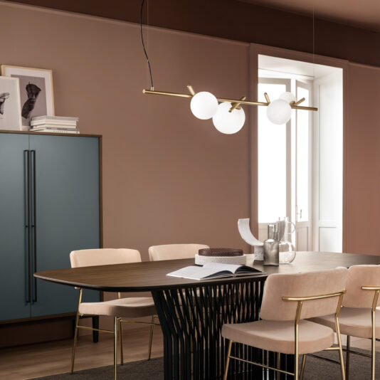 A modern dining room featuring a wooden table with a black base, surrounded by beige chairs adorned with gold accents. Above the table hangs the Contemporary Murano Glass Horizontal Chandelier, showcasing elegant white globes. Against the beige wall, a teal cabinet and framed artwork are displayed. Natural light floods the room through a large window.