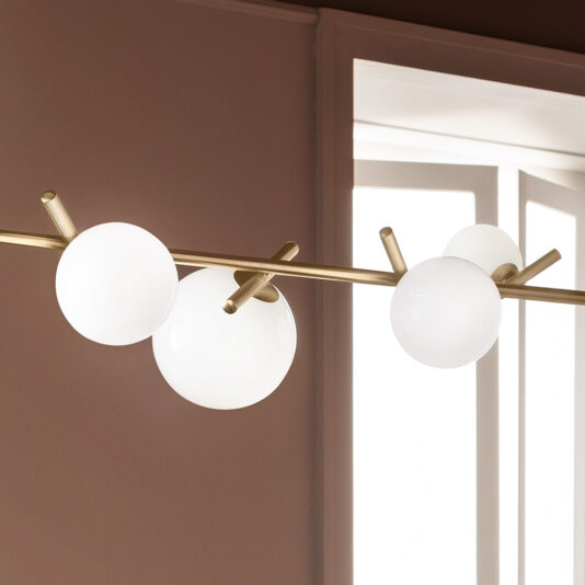 The Contemporary Murano Glass Horizontal Chandelier showcases multiple frosted globes elegantly suspended from a sleek, horizontal golden rod, creating the effect of floating orbs. The scene is complemented by soft natural light filtering through a window framed by beige walls in the background.