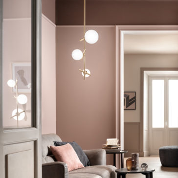 A modern, softly-lit living room with pale pink and taupe walls. A grey sofa with pink and black cushions sits below a Contemporary Murano Glass Vertical Pendant Light featuring two spherical white bulbs. A small round coffee table holds a glass, a candle, and an open book.
