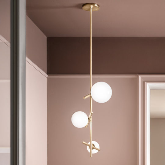 A stunning *Contemporary Murano Glass Vertical Pendant Light* hangs gracefully from the ceiling, featuring a brass fixture with three arms, each topped with a round, white lampshade. The elegant setting is further enhanced by light pink walls and a glimpse of either a white-framed mirror or doorway in the background.