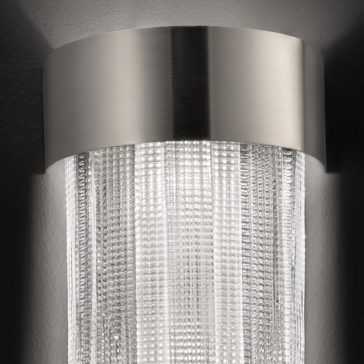 Contemporary Venetian Glass Wall Light A close-up of the Contemporary Venetian Glass Wall Light showcases its cylindrical design. The top is adorned with a metallic silver band, while the lower part features textured Venetian glass panels that create a stunning shimmering effect. The light behind the glass beautifully illuminates the intricate patterns.
