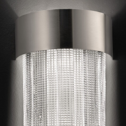 A close-up of the Contemporary Venetian Glass Wall Light showcases its cylindrical design. The top is adorned with a metallic silver band, while the lower part features textured Venetian glass panels that create a stunning shimmering effect. The light behind the glass beautifully illuminates the intricate patterns.